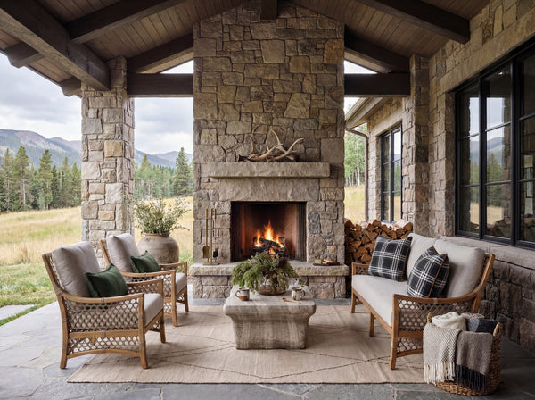 A rustic outdoor patio seating area centered around a stone fireplace, with two woven armchairs, a stone coffee table, stacked firewood, and a view of mountains and trees in the background.