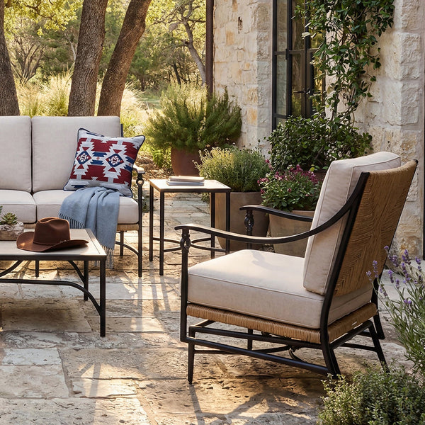 Outdoor patio furniture set arranged on a stone patio between stone buildings, featuring a beige sofa with patterned pillows, two matching armchairs, a rectangular coffee table, and small side tables, surrounded by trees and potted plants.