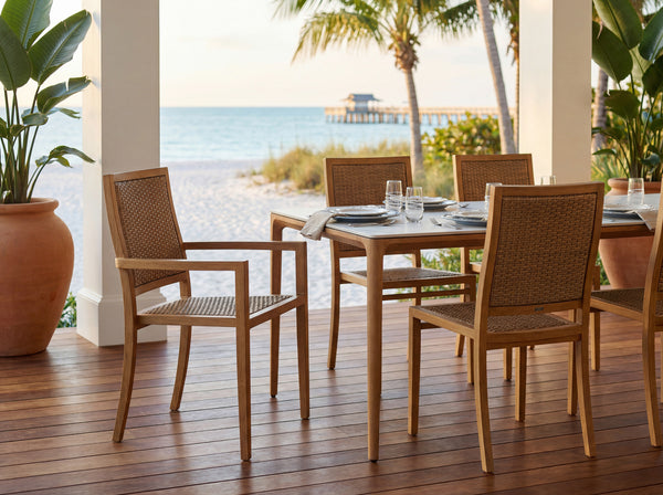 Outdoor dining set with a rectangular wooden table and four woven-back chairs on a wooden deck, overlooking a beach and ocean view.