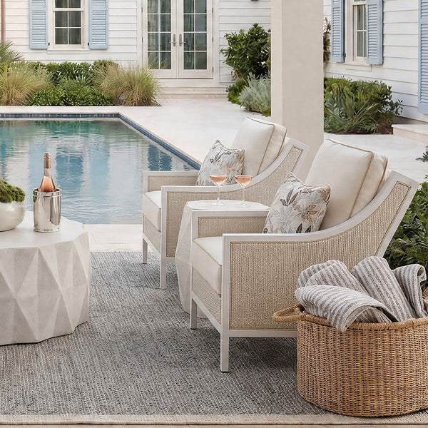 Outdoor patio furniture set arranged on a covered porch beside a swimming pool, featuring a light beige sofa, two matching armchairs, a round geometric coffee table, and a woven basket with folded towels. The background shows a white house with blue shutters and landscaping.